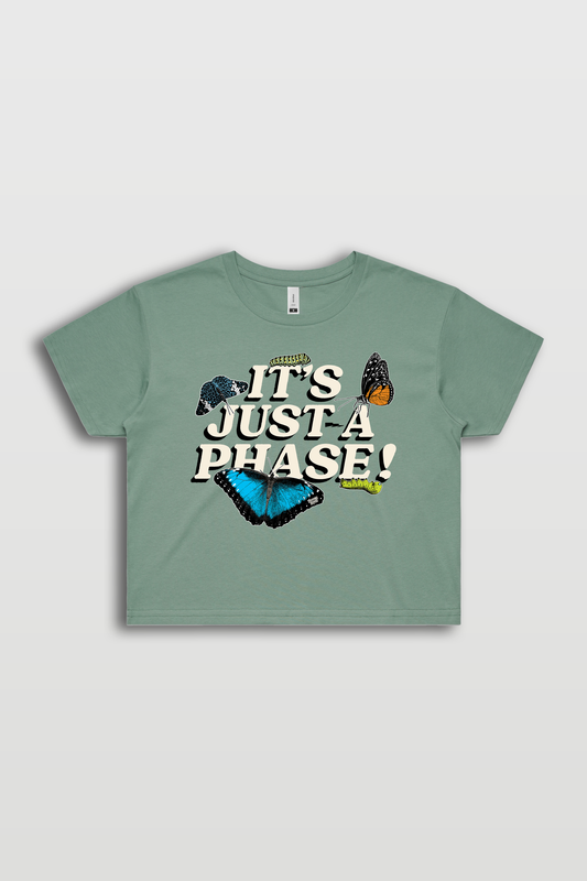 The image displays a stylish and relaxed-fit Street Crop Tee in sage green, featuring a captivating design with butterflies and the phrase "It's just a phase" placed prominently at the center. The tee exudes streetwear vibes with its short body, dropped shoulders, and boxy fit. It's a high-quality 100% Cotton garment, pre-shrunk to maintain its shape. Please note that this tee is created specifically when ordered to minimize environmental impact and ensure humane production.